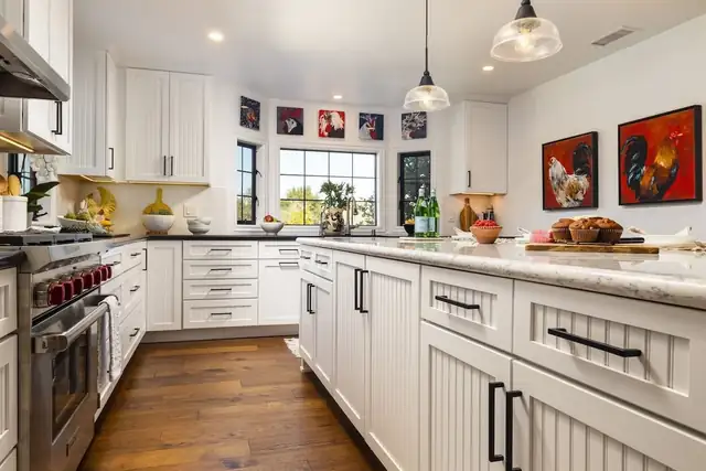 White kitchen by ELITE Remodels with beadboard cabinet panels, black pulls, a professional range, wood floors and red art prints
