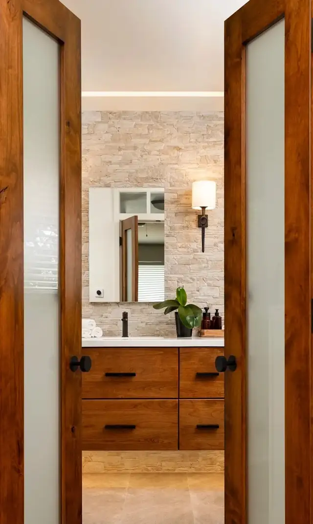Santa Barbara bathroom seen through frosted glass double doors, with stacked stone wall, wood vanity and wall sconces