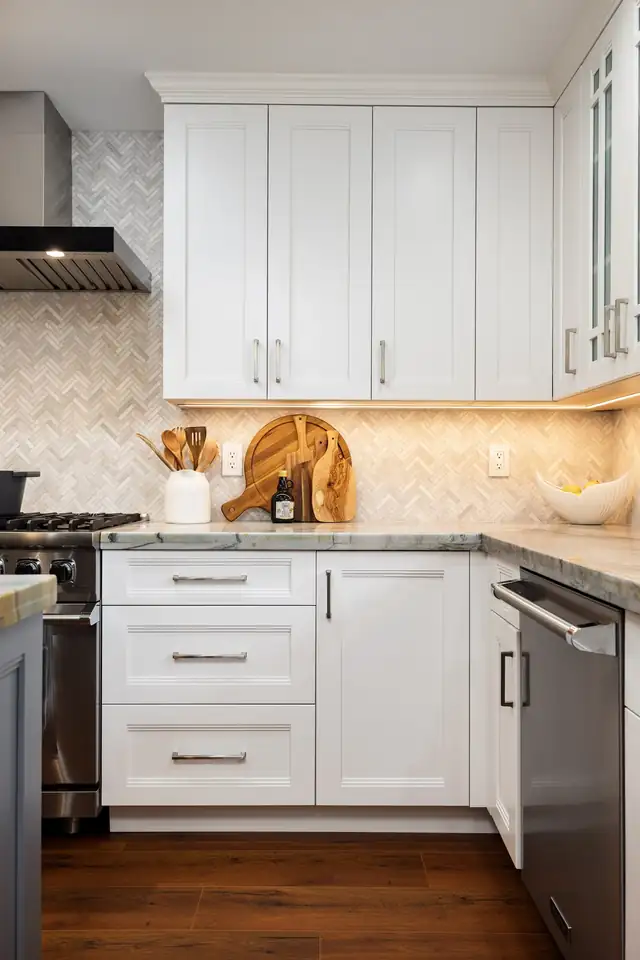 Kitchen corner with white shaker cabinets, glass-front uppers, a cream herringbone backsplash and warm under-cabinet lighting